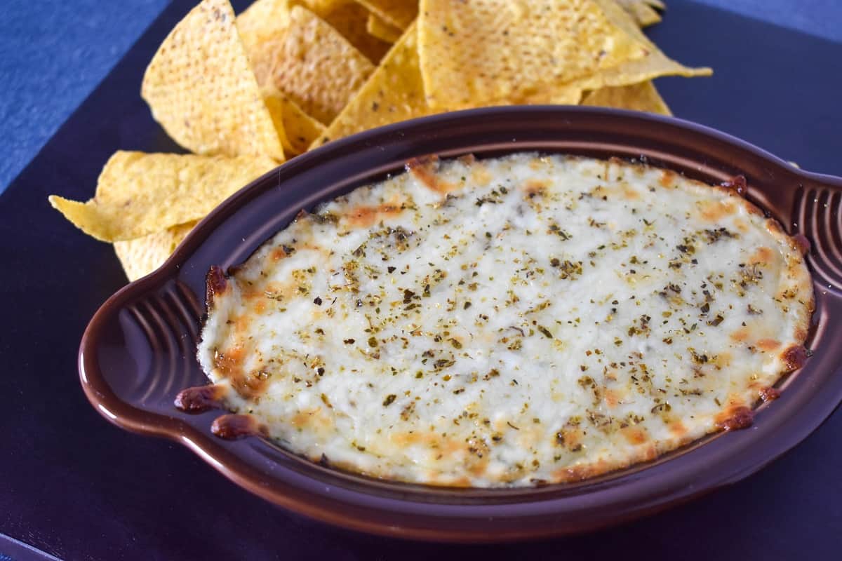 Hot spinach dip served in a brown bowl with corn tortilla chips in the background.