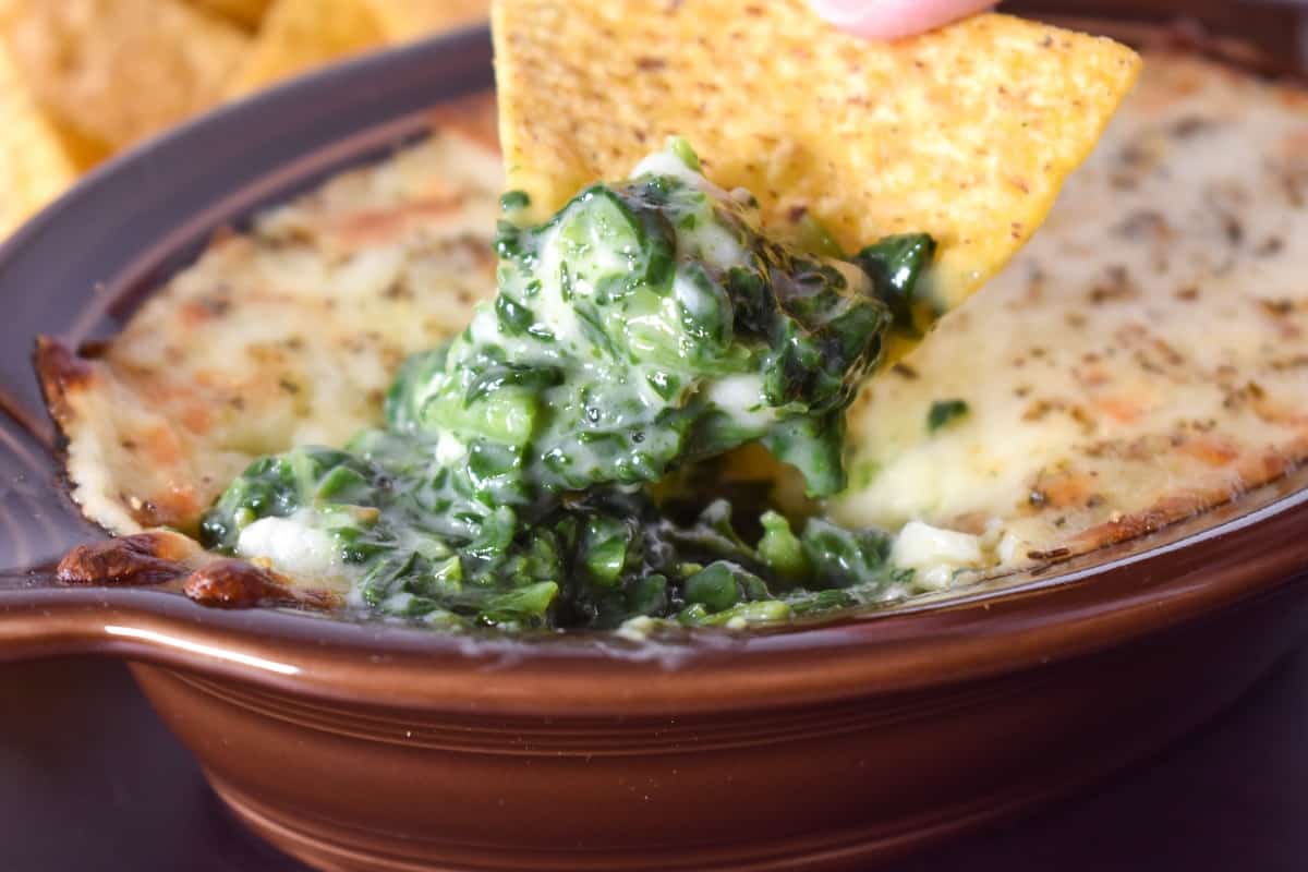 Creamy spinach and cheese being scooped out of a brown crock with a corn tortilla chip.