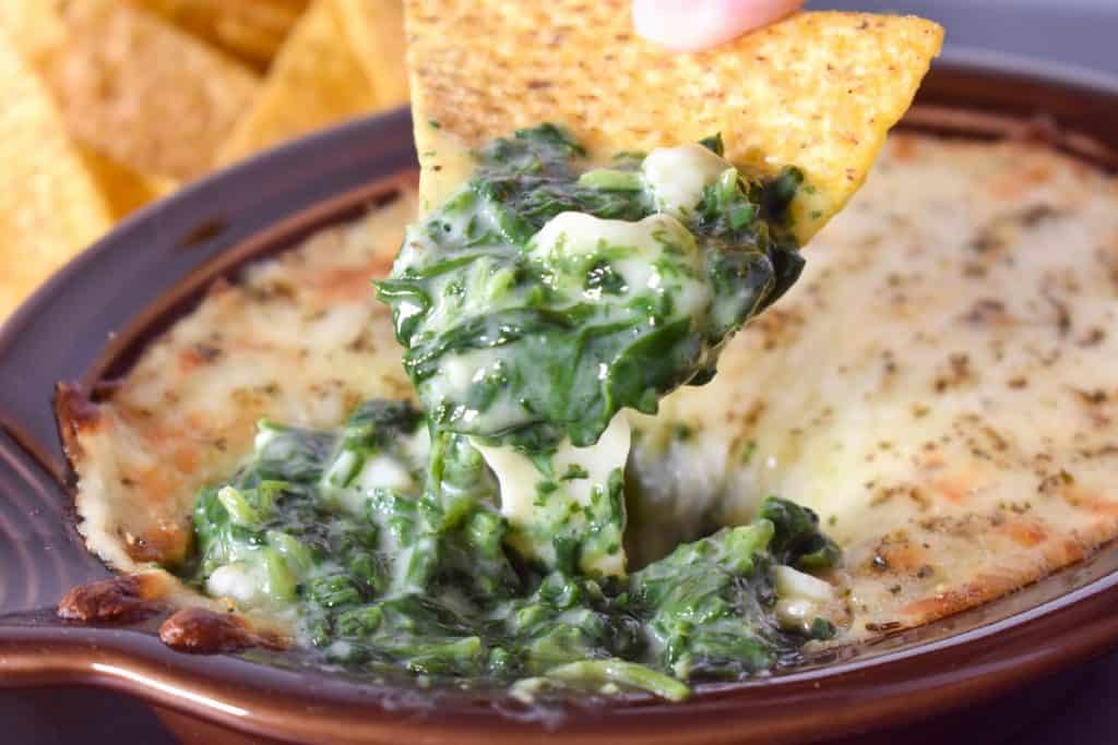 Creamy spinach and cheese being scooped out of a brown crock with a corn tortilla chip.