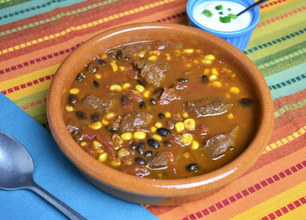 Southwestern Steak Soup with chunks of beef, black beans, tomatoes,and corn in a beef broth, served in a red clay bowl with sour cream on the side.