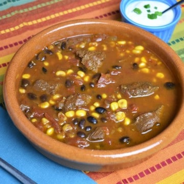 Southwestern Steak Soup with chunks of beef, black beans, tomatoes,and corn in a beef broth, served in a red clay bowl with sour cream on the side.