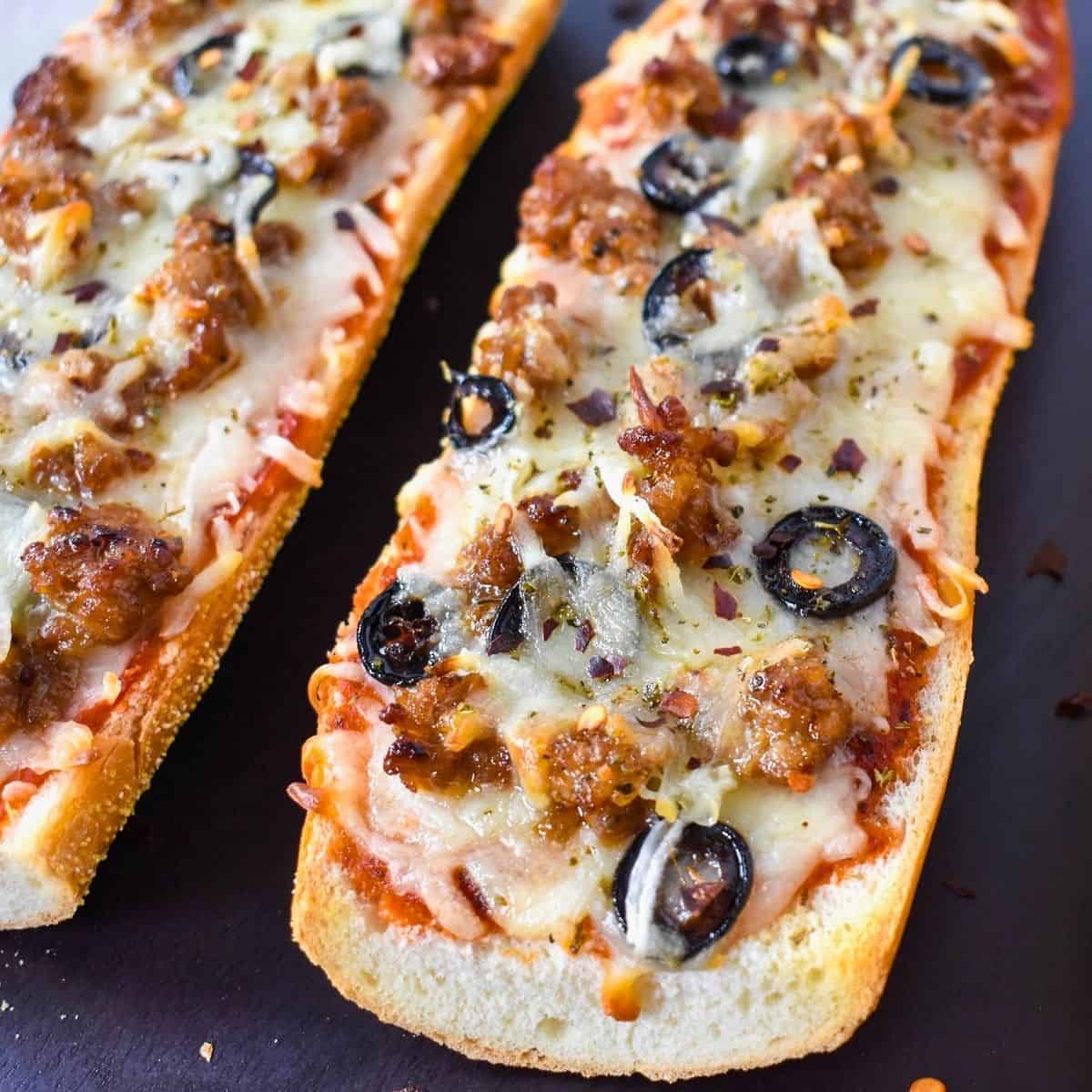 French bread pizza with sausage, black olives and thin onions on a black cutting board.