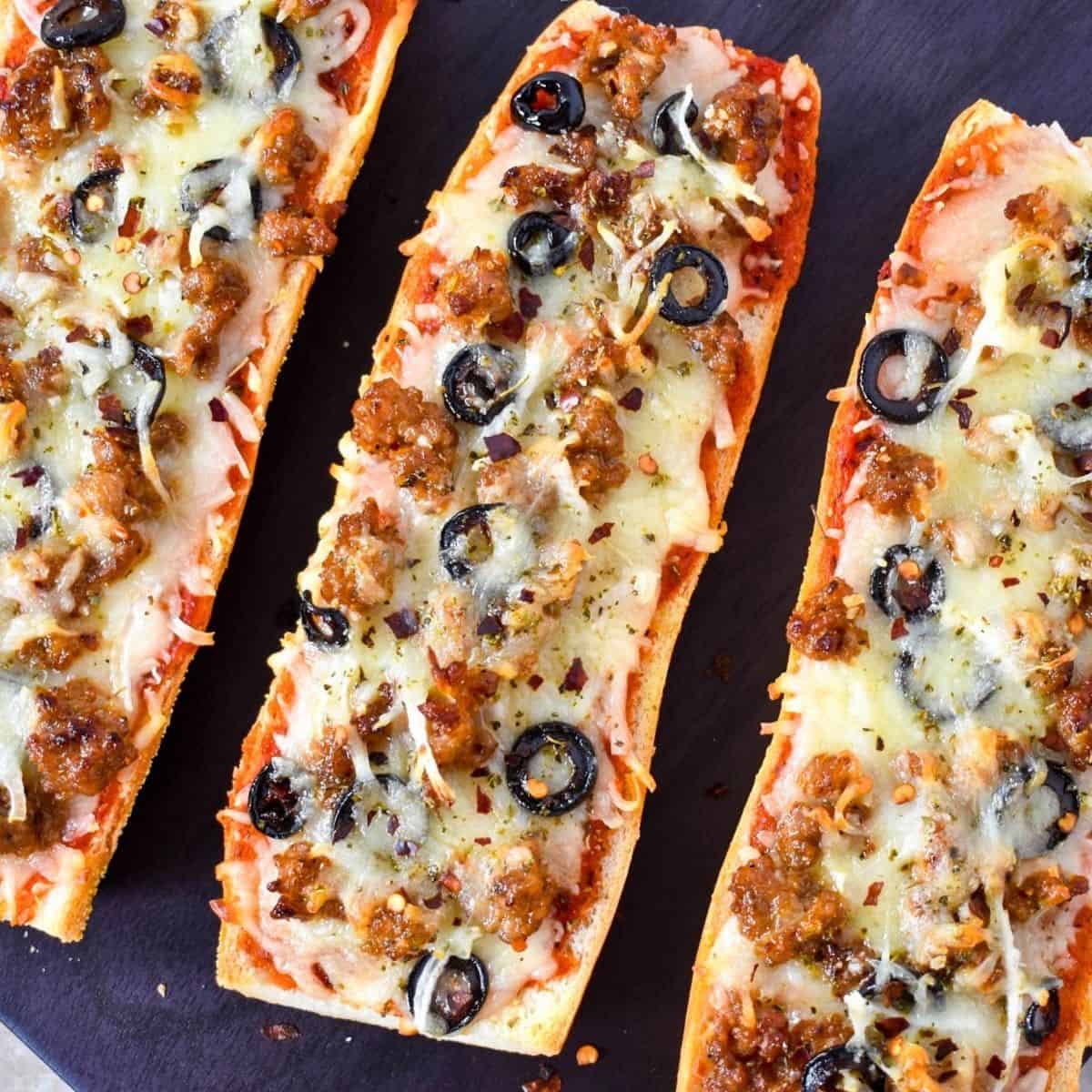 French bread pizza with sausage, black olives and thin onions on a black cutting board.