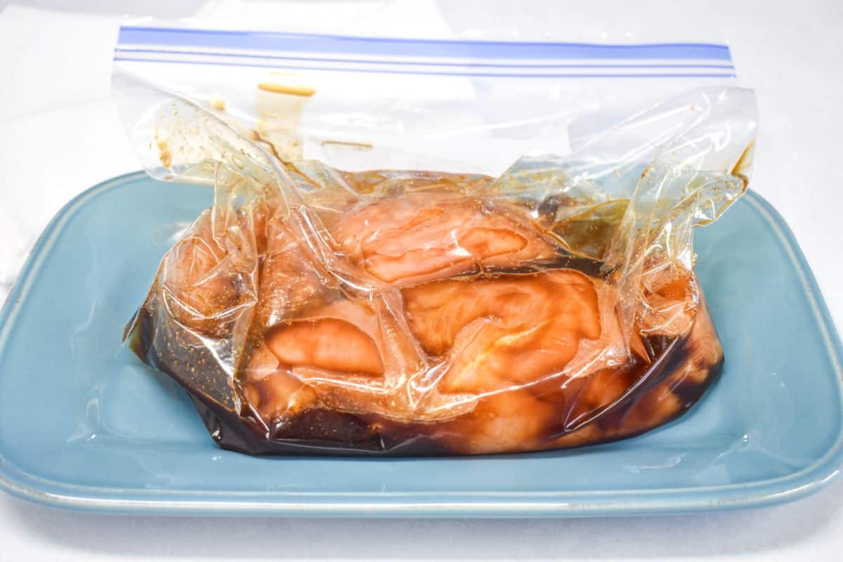 Chicken breasts marinating in soy-based marinade inside a sealed plastic bag on a plate.