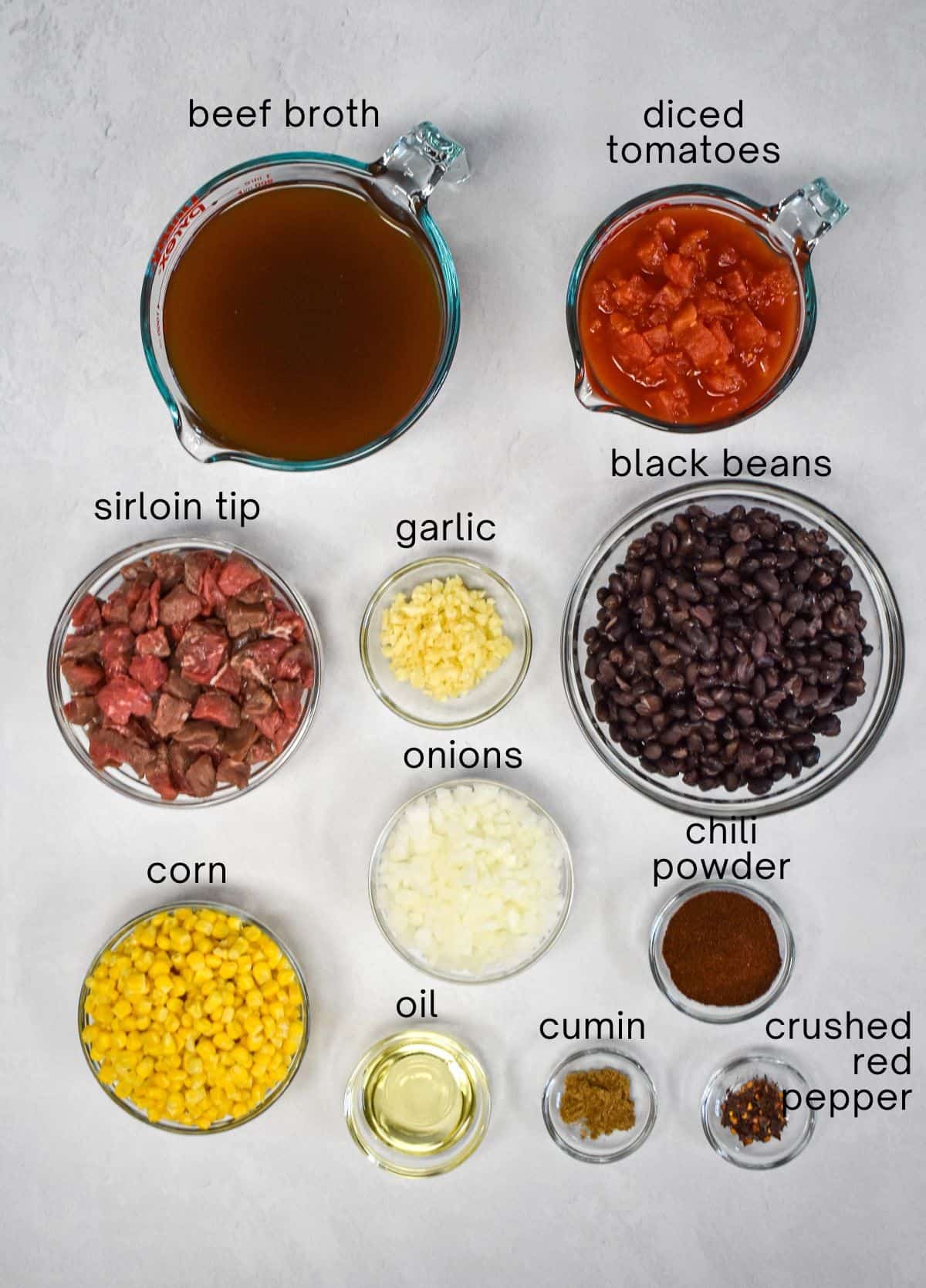 The ingredients for the soup arranged in glass bowls on a white table with each labeled in small, black letters.