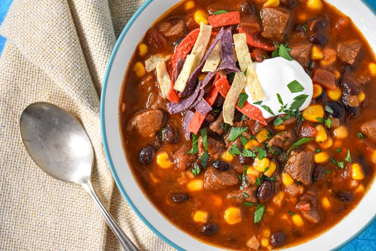 A close up of the southwestern steak soup served in a white bowl with a light blue rim and garnished with tortilla strips, sour cream, and parsley.