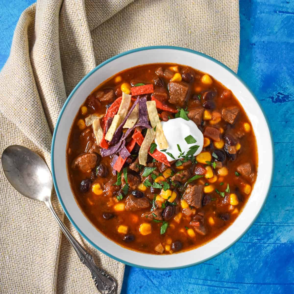 The southwestern steak soup served in a white bowl with a light blue rim set with a beige linen on a blue table.