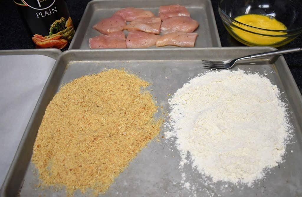 Breading Station, with flour and breadcrumbs on a pan, a beaten egg in a bowl and chicken breast cutlets in the background