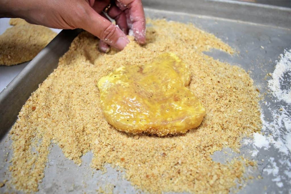 Breading, a chicken breast cutlet that was dredged in flour, dipped in egg sits on a bead of breadcrumbs