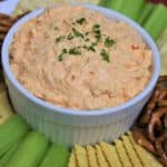 Cold Buffalo Chicken Dip served in a white bowl with assorted crackers, celery sticks and pretzels arranged on the plate.