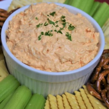 Cold Buffalo Chicken Dip served in a white bowl with assorted crackers, celery sticks and pretzels arranged on the plate.