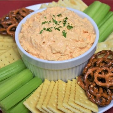 Cold Buffalo Chicken Dip served in a white bowl with assorted crackers, celery sticks and pretzels arranged on the plate.