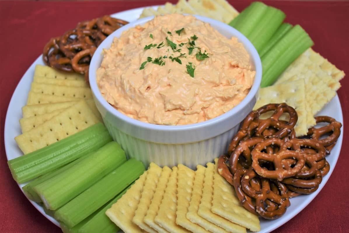 Cold Buffalo Chicken Dip served in a white bowl with assorted crackers, celery sticks and pretzels arranged on the plate.