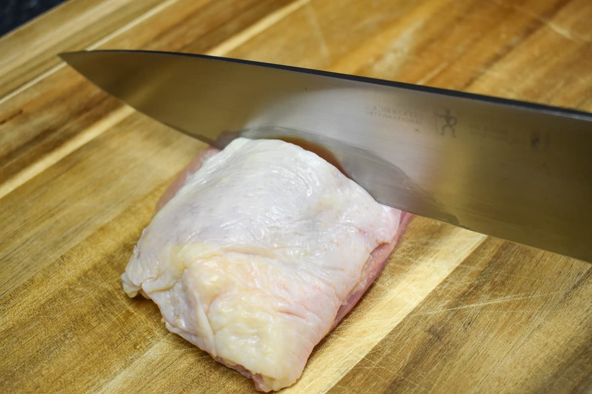 A knife going through a chicken thigh on a wood cutting board.
