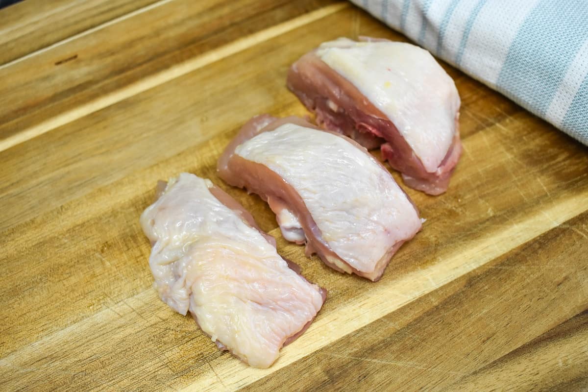 A chicken thigh cut into three pieces displayed on a wood cutting board.