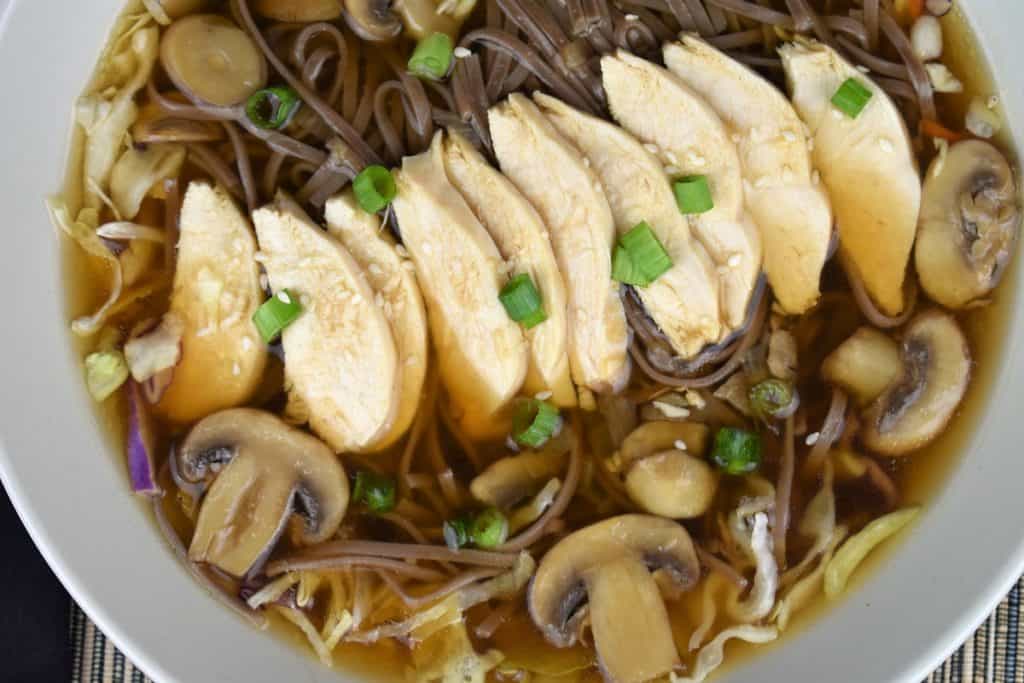 A close up of soba noodle chicken soup with mushrooms, green onions and sesame seeds.