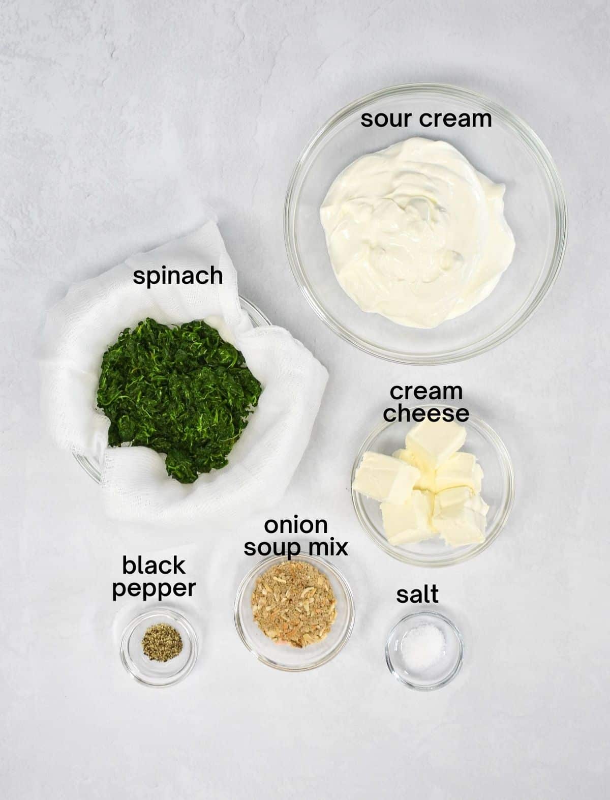 The ingredients for the dip arranged in glass bowls on a white table. Each ingredients has the name above it in black letters.