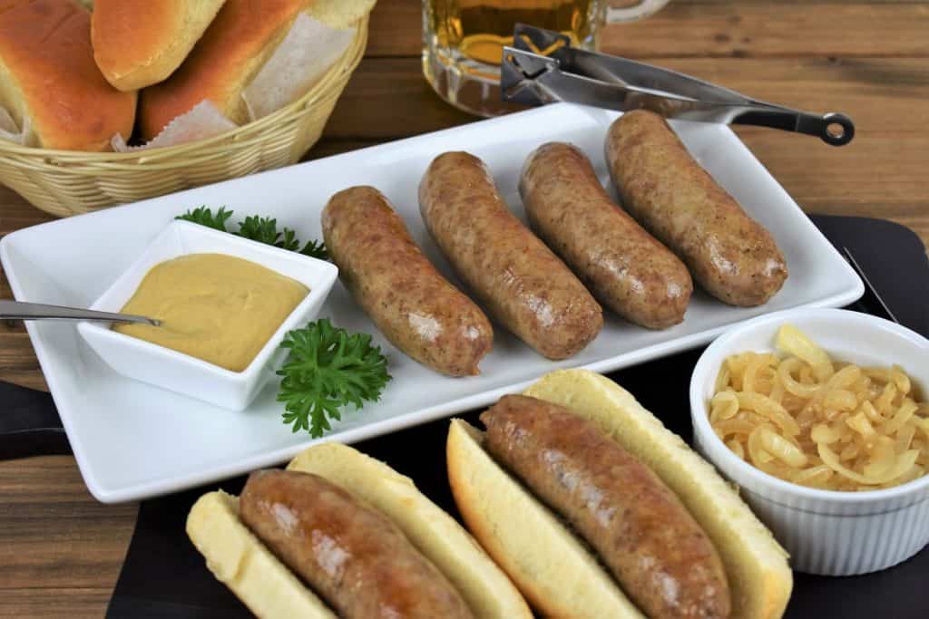 Beer brats, sausages arranged on a white platter with two of the brats served on buns with a small ramekin of mustard and a ramekin of cooked, sliced onions on the side