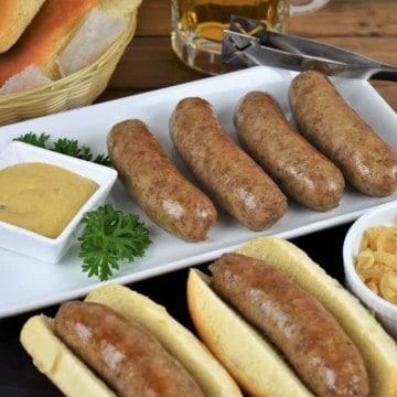 Beer brats, sausages arranged on a white platter with two of the brats served on buns with a small ramekin of mustard and a ramekin of cooked, sliced onions on the side