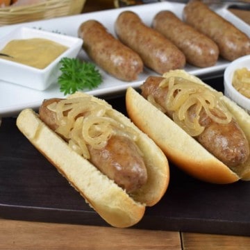 Beer brats, sausages arranged on a white platter with two of the brats served on buns and topped with cooked onions