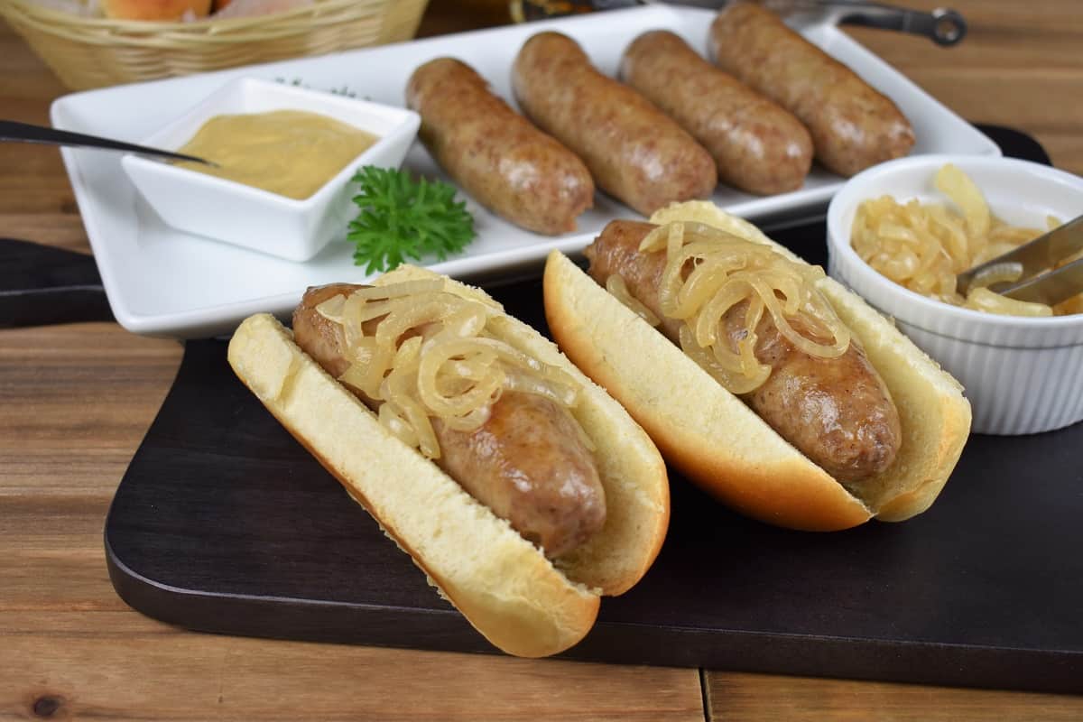 Beer brats, sausages arranged on a white platter with two of the brats served on buns and topped with cooked onions
