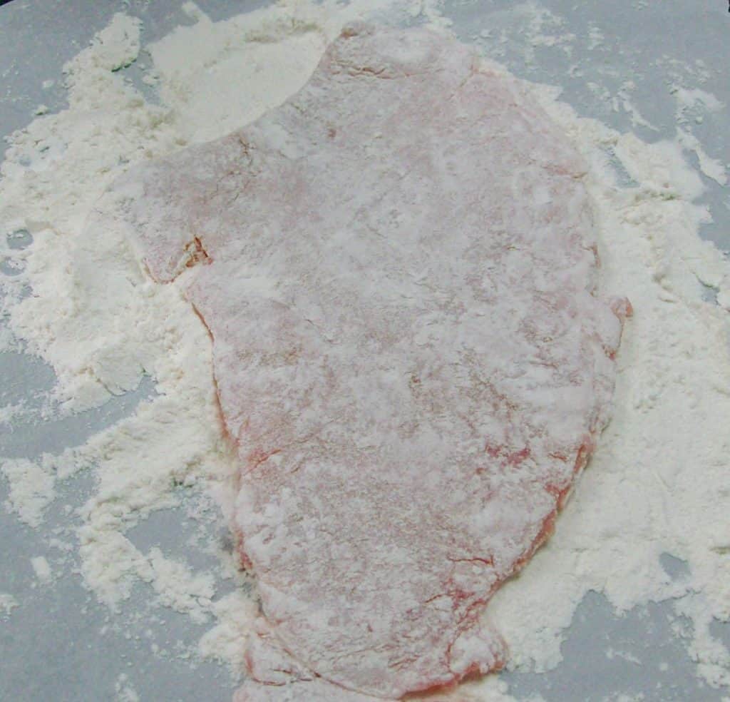 A thin piece of pork dredged with flour on a metal baking sheet.