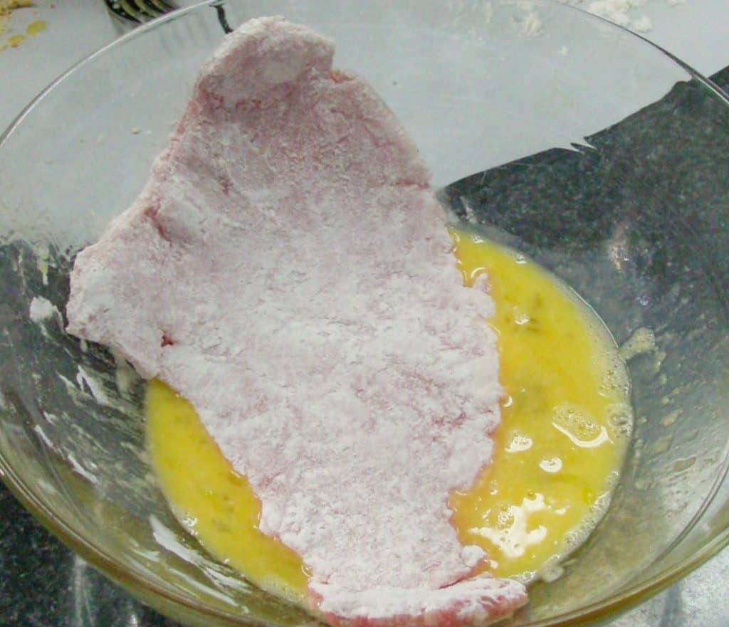 A thin piece of pork that's dredged in flour being dipped into an egg bowl.