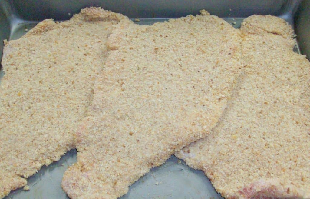 Uncooked breaded pork steaks displayed on a metal baking sheet.