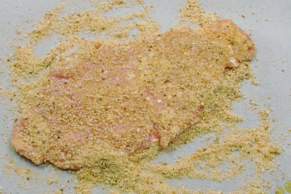 A thin piece of pork being breaded on a metal baking sheet.