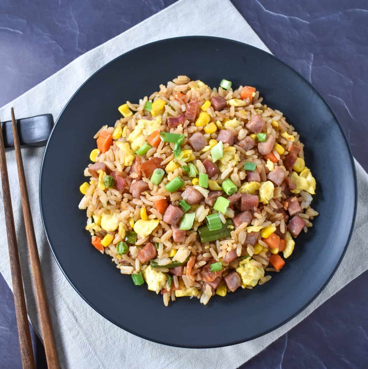 The ham fried rice served in a black plate and placed on a beige linen on a black table. There are chopsticks to the left hand side.