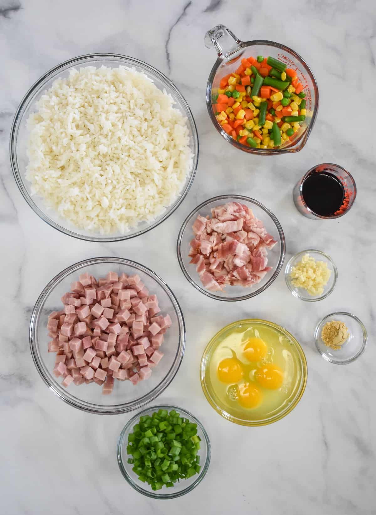 The ingredients for the ham fried rice prepped and separated in glass containers arranged on a white table.
