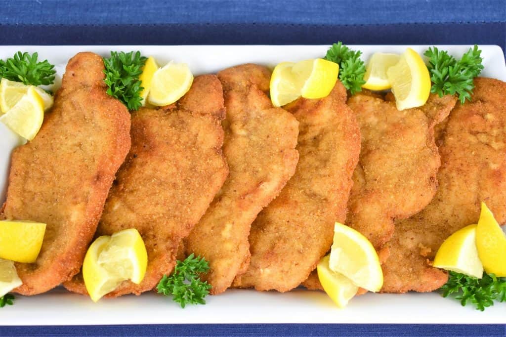 Pork schnitzel arranged on a white platter and garnished with fresh parsley and lemon wedges.