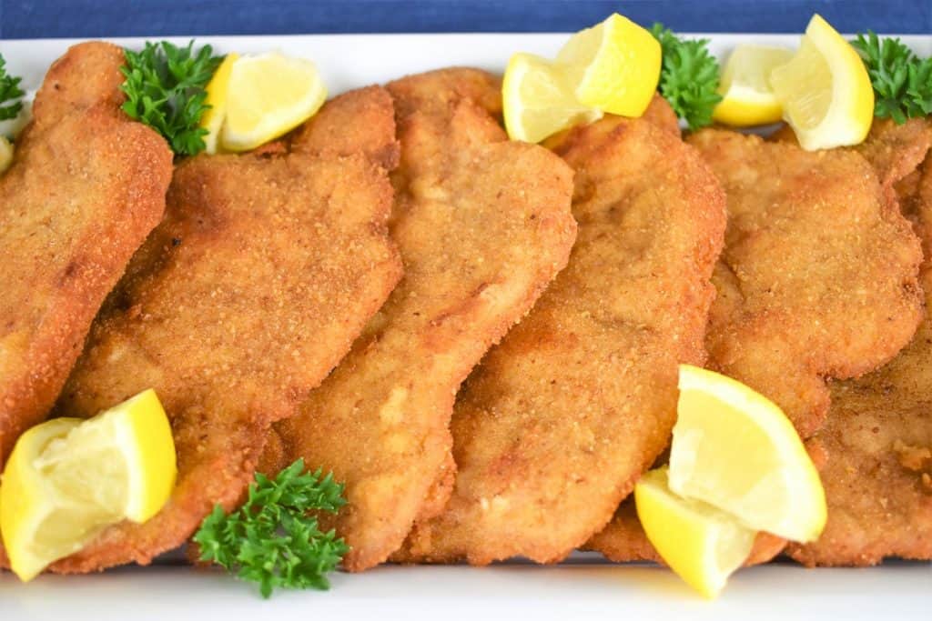 Pork Schnitzel arranged on a white platter and garnished with fresh parsley and lemon wedges.