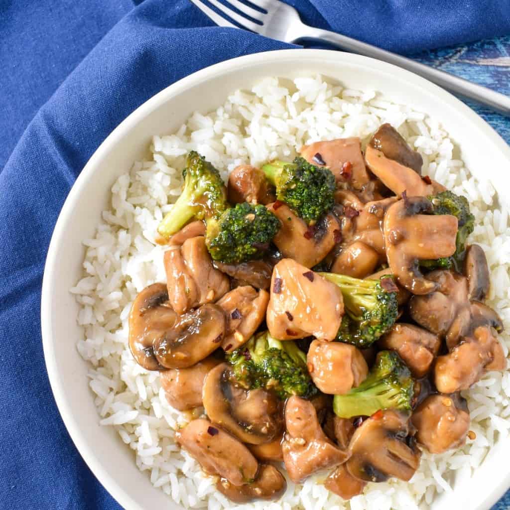 A close up of the chicken and broccoli stir fry served on a bed of white rice in a white bowl. There is a blue linen and a fork off the the top right side.
