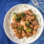 An image of the chicken broccoli stir fry served on a bed of white rice, served in a white bowl with a blue linen and set on a blue table.