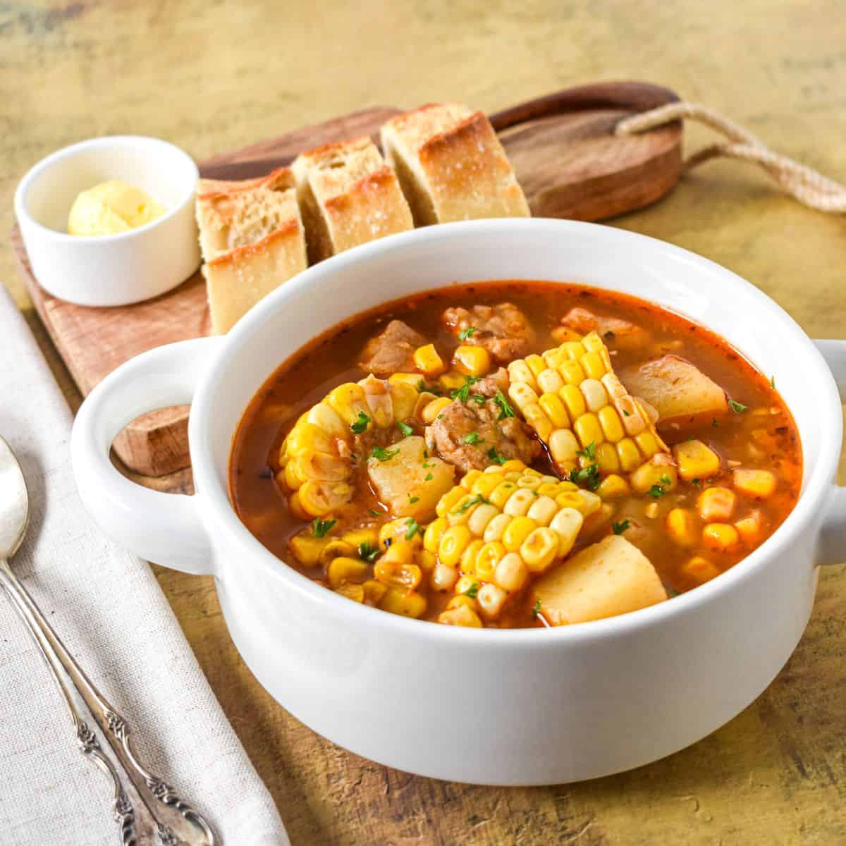 Labeled ingredients for guiso de maíz arranged on a white surface, including pork, corn, potatoes, broth, onion, garlic, and seasonings.
