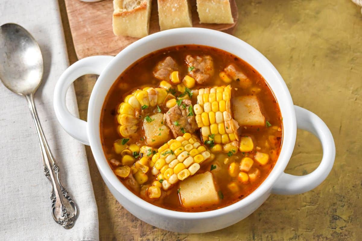 Cuban corn stew in a white bowl with chunks of pork, corn, and potatoes in broth, served with bread.