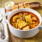 Cuban corn stew in a white bowl, filled with corn cobs, potatoes, and pork in broth, with bread and butter on a wooden board.
