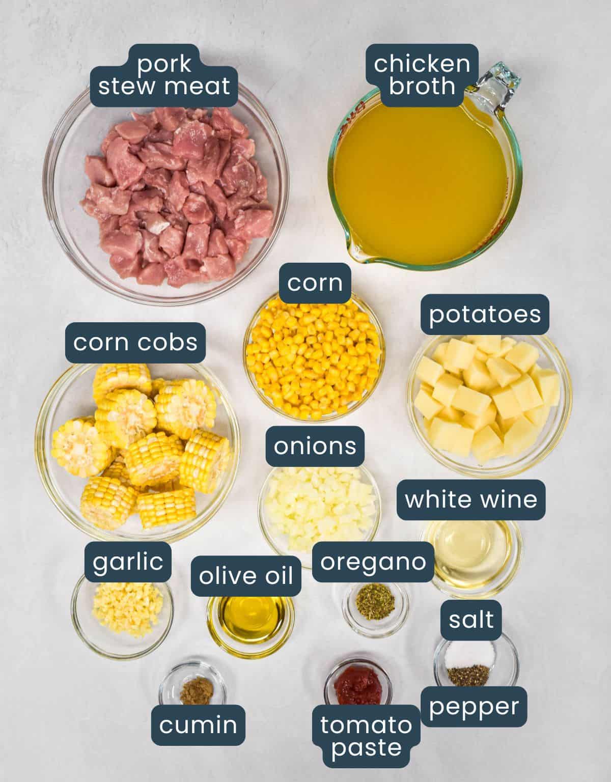 Labeled ingredients for dish arranged on a white surface, including pork, corn, potatoes, broth, onion, garlic, and seasonings.