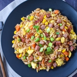 The ham fried rice served in a black plate and placed on a beige linen on a black table. There are chopsticks to the left hand side.