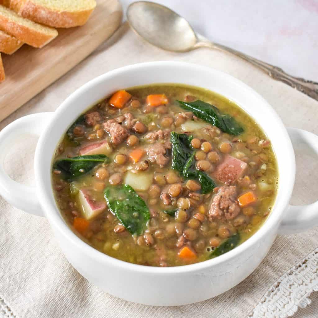 The Italian sausage lentil soup served in a white bowl set on a linen with a spoon and sliced bread in the background.