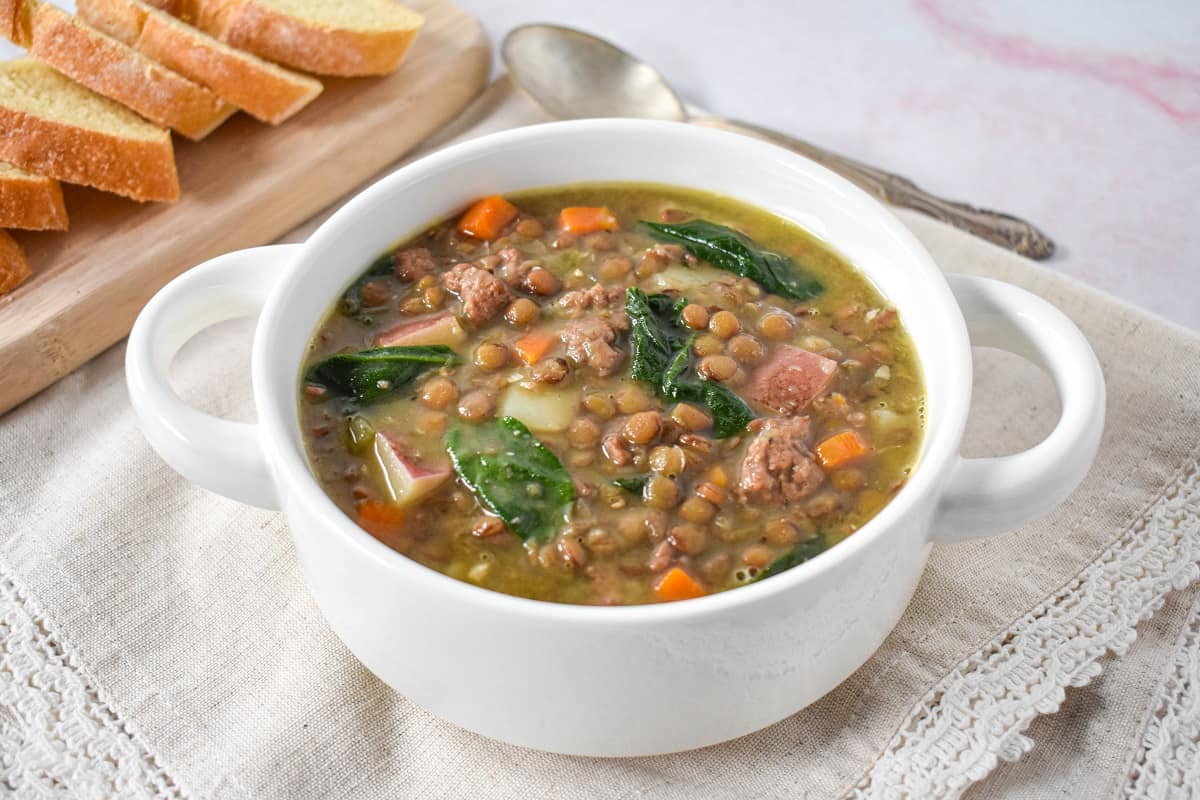 The Italian sausage lentil soup served in a white bowl set on a linen with a spoon and sliced bread in the background.