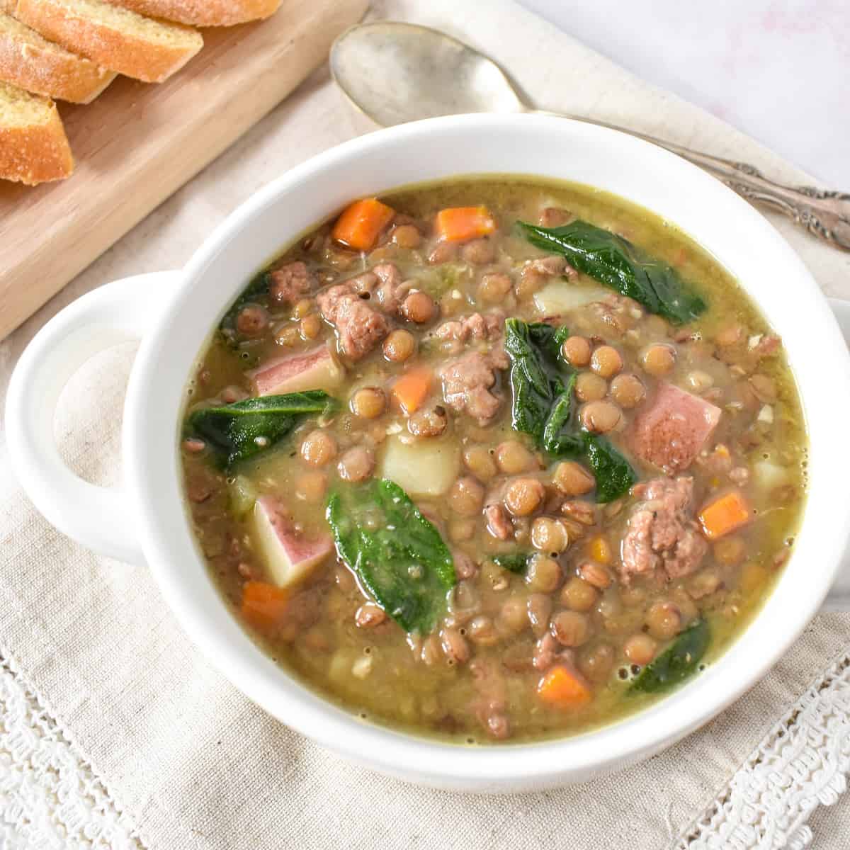 The Italian sausage lentil soup served in a white bowl set on a linen with a spoon and sliced bread in the background.