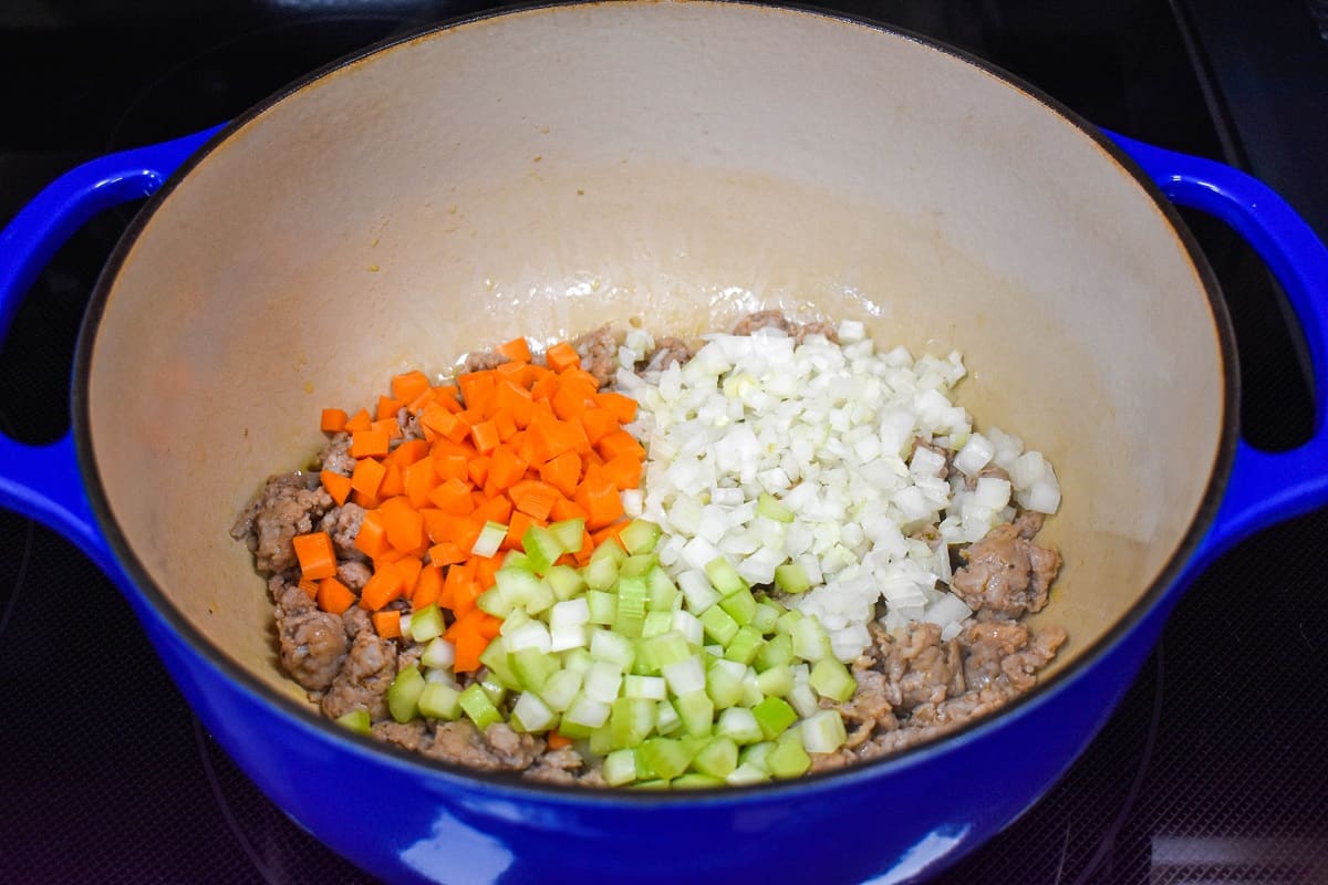 Onions, carrots, and celery over browned sausage in a large, blue and white pot.