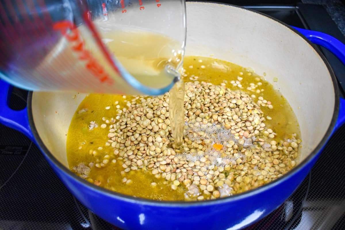 Chicken broth being added to the lentils and other ingredients in a large, blue and white pot.