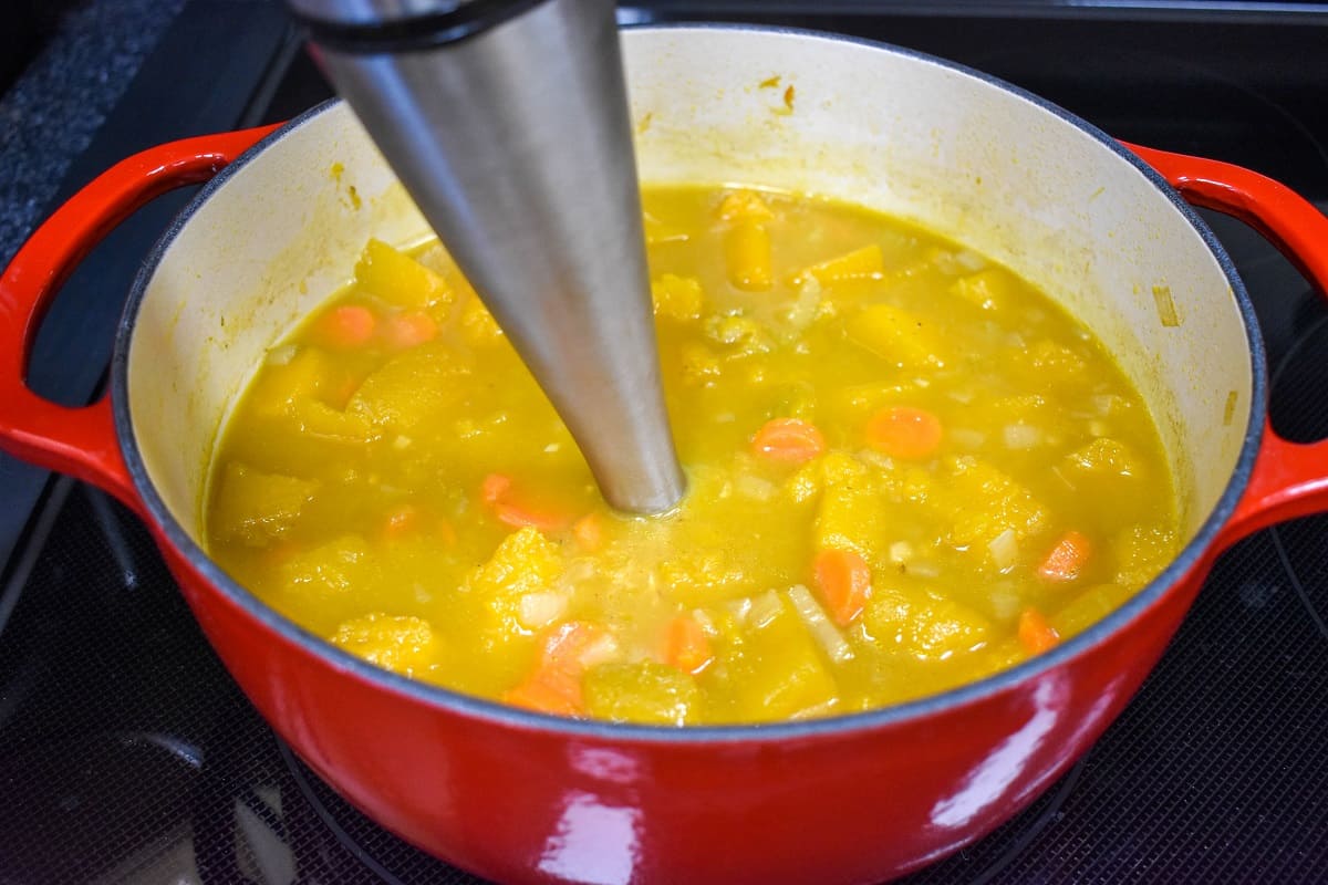 The pumpkin ginger soup being processed with an immersion blender in a red pot.