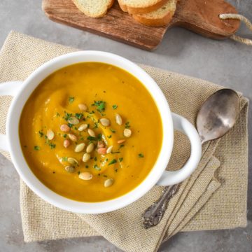 The pumpkin ginger soup garnished with pumpkin seeds and chopped parsley in a white bowl set on a beige linen. In the background there is a small wood cutting board with sliced bread and a spoon to the right side.