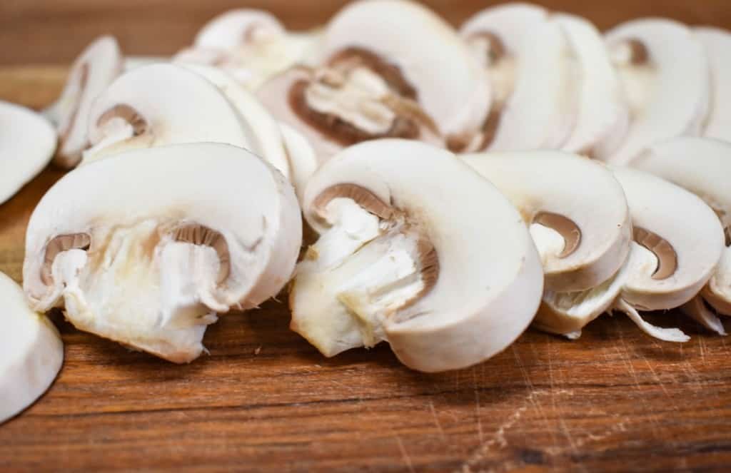 A close up image of sliced white button mushrooms on a wood cutting board.
