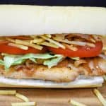 A Cuban chicken sandwich displayed on a wood cutting board. A thin chicken breast on Cuban bread topped with shredded lettuce, sliced tomatoes and potato sticks.