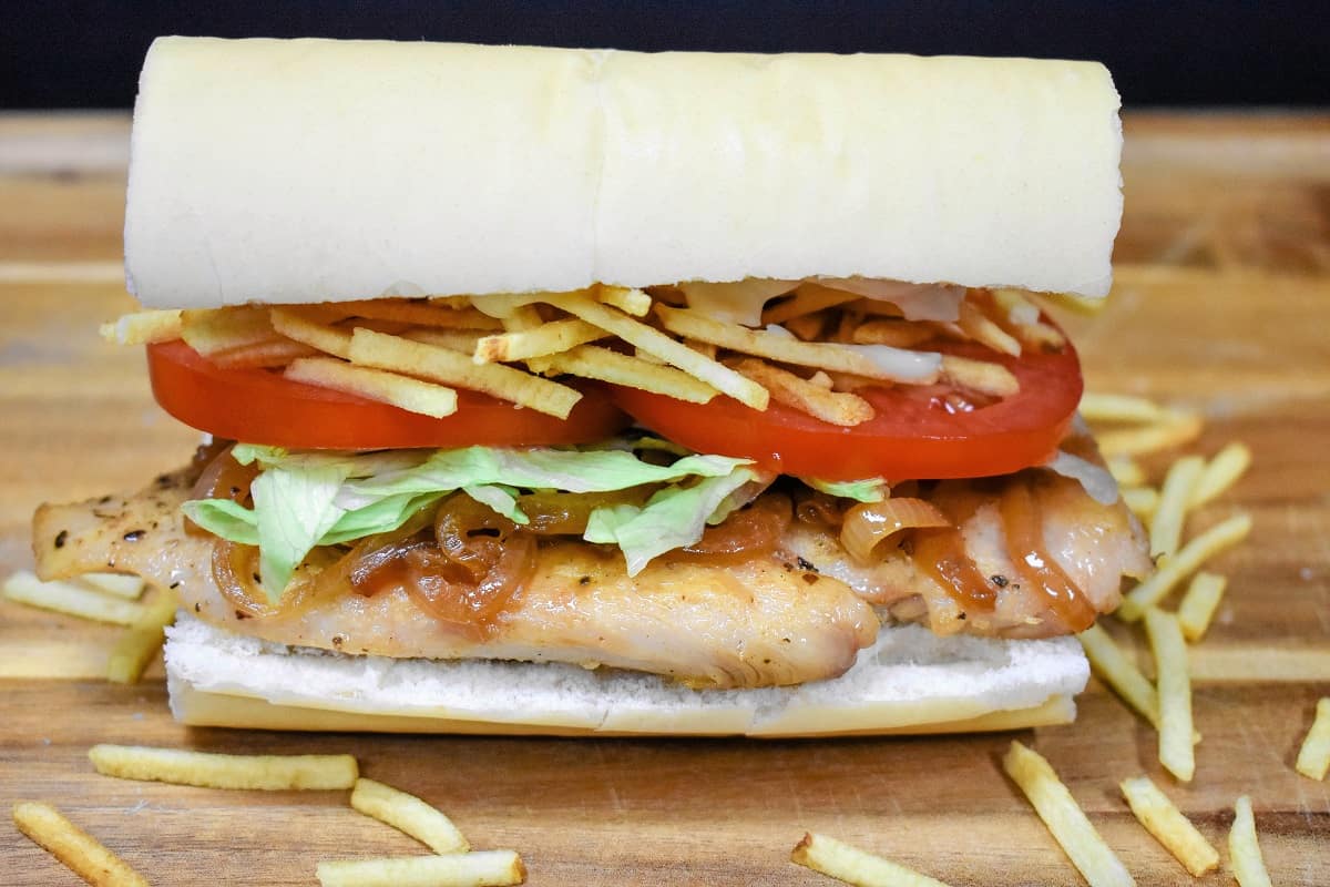 A Cuban chicken sandwich displayed on a wood cutting board. A thin chicken breast on Cuban bread topped with shredded lettuce, sliced tomatoes and potato sticks.